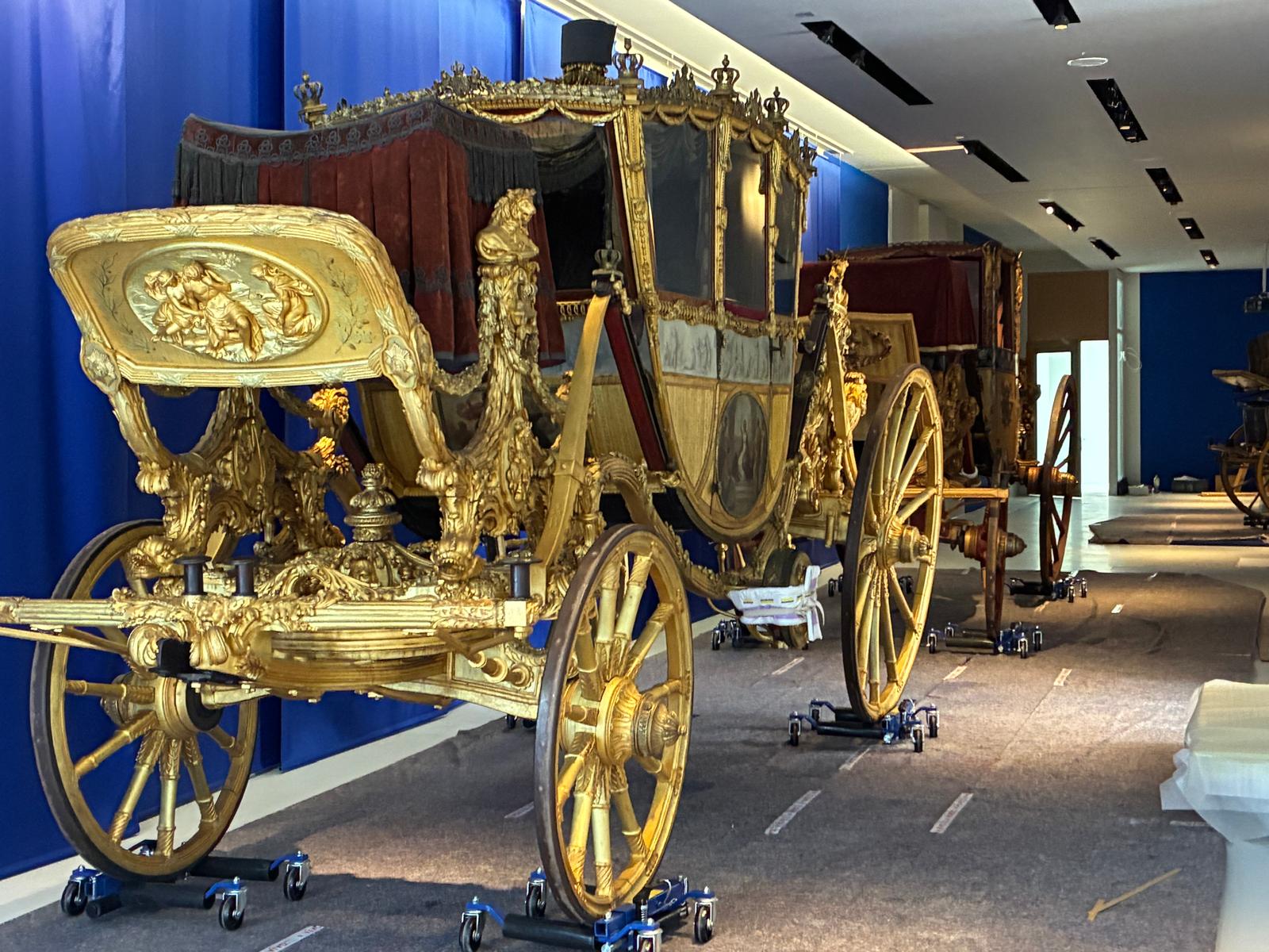 Ein Foto einer goldenen Kutsche vor einer blauen Wand. Unter den Rädern stehen Rollbretter.